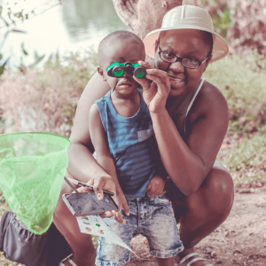 Toddler and mother crouched in front of body of water with binoculars and a net ready to explore!